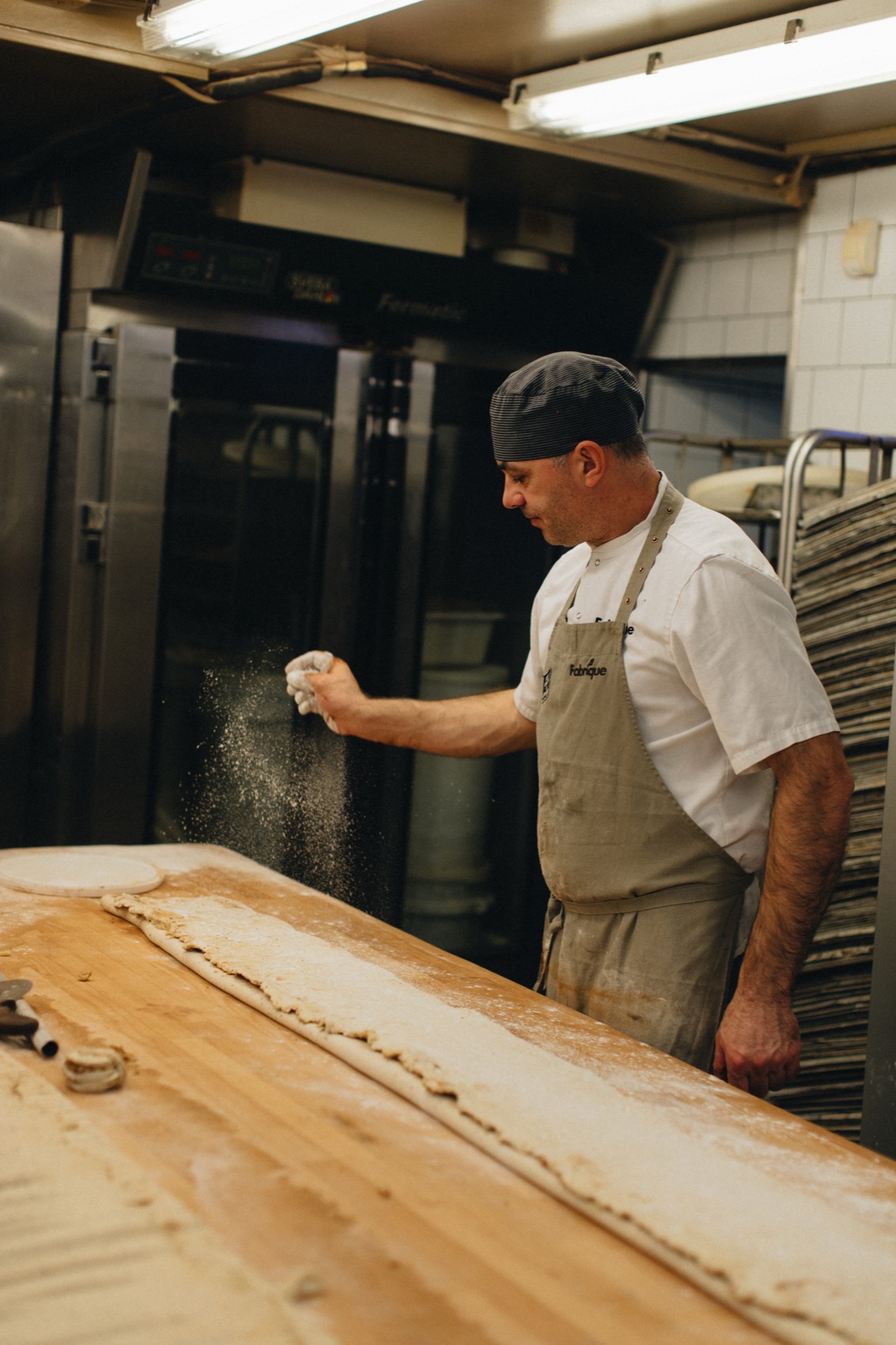 A baker in a Fabrique apron dusting flour over a long piece of dough in the bakery.